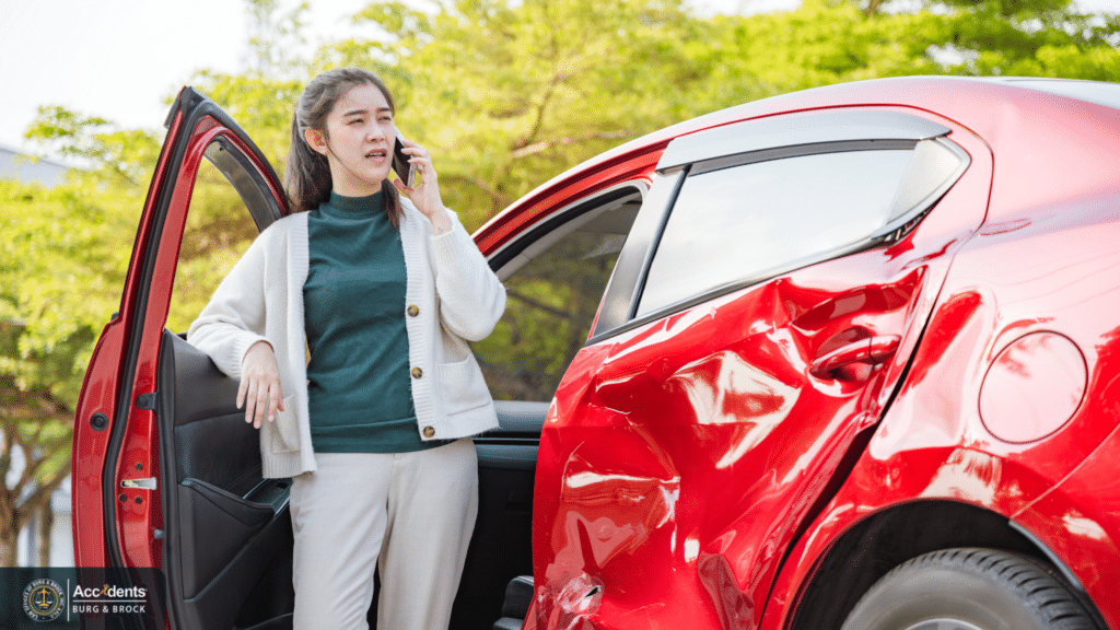 A woman stands beside her open car door making a phone call after a collision, with the rear quarter panel of a red car badly dented.