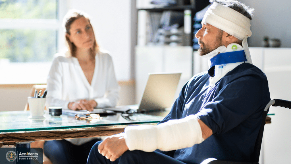 Injured man in a wheelchair with a bandaged head, neck brace and arm cast, sitting across a desk from a woman taking notes at a laptop
